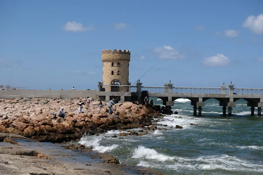 Waves breaking on the rocks below a stone tower and pier in Alexandria, shown as a matted print